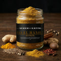 A glass jar filled with Ras el Hanout spice blend sits on a dark wooden surface surrounded by whole spices, ginger root, cinnamon, and chili powder. The jar has a black label with white and gold text reading 'NEVER NOT EATING' and 'RAS EL HANOUT'.
