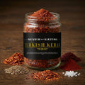 A clear glass jar filled with red Turkish kebab seasoning blend sits on a dark wooden surface, surrounded by loose piles of red chili flakes, cumin seeds, and coarse salt. The jar has a black label reading 'NEVER NOT EATING TURKISH KEBAB'.