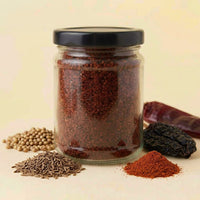 A clear glass jar filled with reddish-brown ground spice or chili powder, with a black lid, photographed on a warm cream background. Small piles of whole spices and dried red chilies are arranged around the base of the jar.