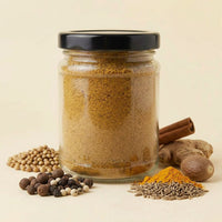 A clear glass jar filled with a brown spice blend and topped with a black lid, styled on a light beige background with whole spices and ingredients including cinnamon sticks, turmeric powder, coriander seeds, peppercorns, and ginger roots.