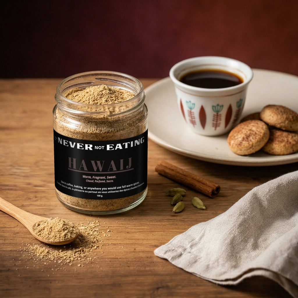 Jar of Never Not Eating Hawaij spice blend beside a cup of black coffee and biscuits, with cinnamon, cardamom and a wooden spoonful of seasoning.