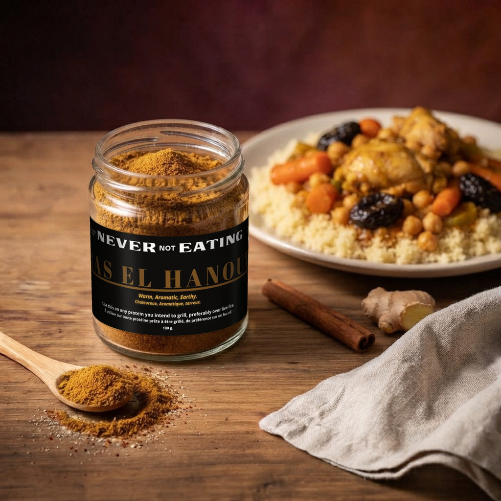 Jar of Never Not Eating Ras el Hanout beside chicken, carrots, chickpeas and dried fruit served over couscous, with a wooden spoonful of spice.